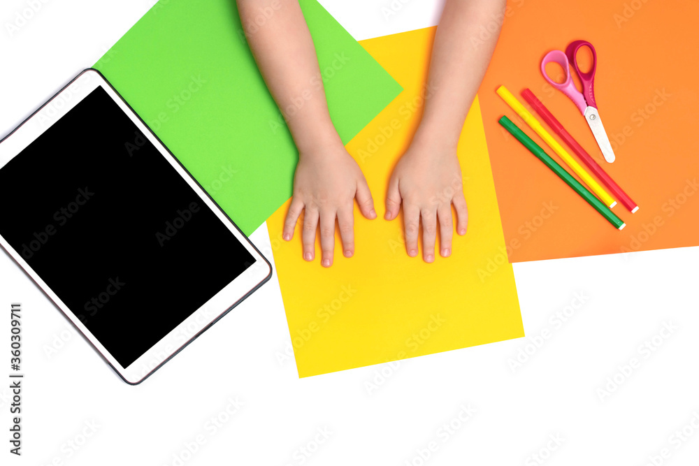 Tablet, multi-colored sheets of paper and the hands of a child on the table. View from above. Online classes on the tablet. Copy space Tablet, multi-colored sheets of paper and the hands of a child on the table. View from above. Online classes on the tablet. Copy space