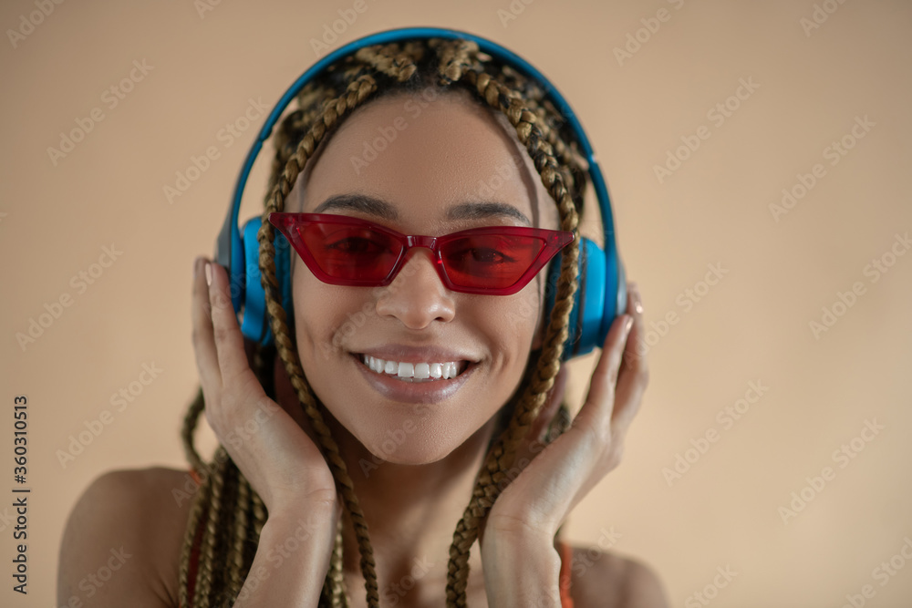 Smiling young African American female in red eyeglasses, touching blue headphones and listening to music