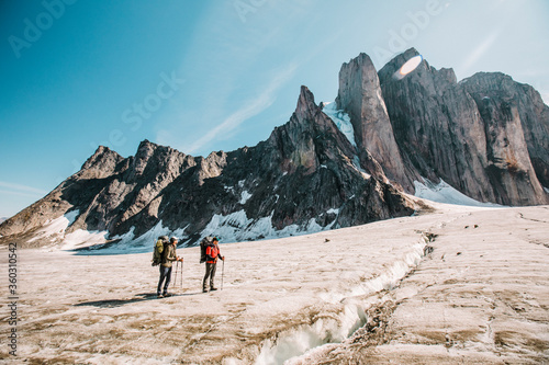 Two explorers standing on glcier below steep mountain summits.