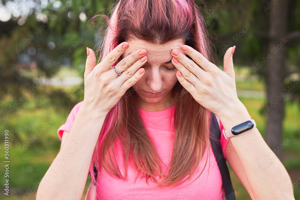 woman have a headache pain outdoors. girl has pink hair and a pink colored shirt. female closed her eyes and holds hands on head. woman have a headache pain outdoors. girl has pink hair and a pink colored shirt. female closed her eyes and holds hands on head.