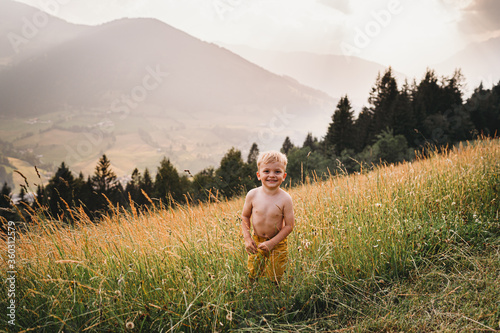 White male toddler smiling standing topless in the grass in mountains