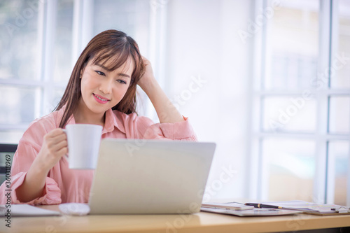 Beautiful Asian woman with long hair Wearing a pink shirt holding a white coffee cup and looking at the laptop. Relax drinking coffee in office concept.