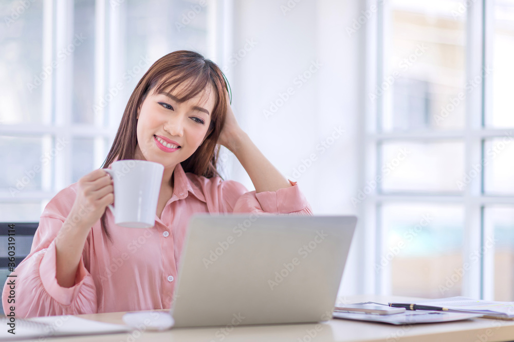 Beautiful Asian woman with long hair Wearing a pink shirt holding a white coffee cup and looking at the laptop. Relax and drinking coffee in office, break concept. Beautiful Asian woman with long hair Wearing a pink shirt holding a white coffee cup and looking at the laptop. Relax and drinking coffee in office, break concept.