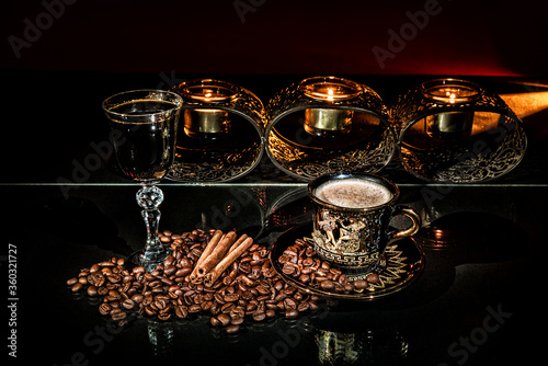 Cup of coffee, with roasted beans and cinnamon sticks around. Glass with black alcoholic drink and behind candle holder with burning candles on the black background. Still life photo. Luxury concept.