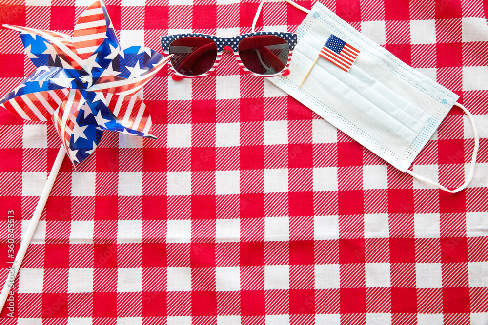 Obraz premium July 4 background. USA flags, sunglasses, and medical mask on red tablecloth. Independence Day of America during coronavirus covid-19 pandemic quarantine. Flat lay, top view, daylight. Copy space text
