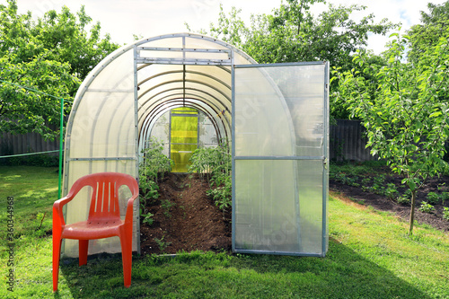 Polycarbonate greenhouse with open door in a well-kept garden. In the evening sunlight, you feel calm and comfort