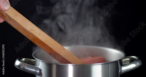 Male hand holding forceps gets boiled sausage from pan.