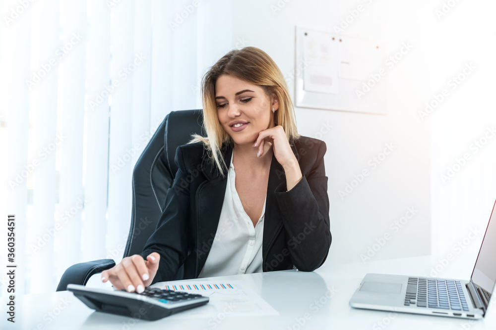 Young confident businesswoman sitting in her bright office while looking at the calculator on office desk. Young confident businesswoman sitting in her bright office while looking at the calculator on office desk.