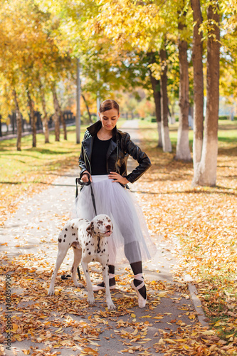 Ballerina with Dalmatian dog in the Park. Woman ballerina in a white ballet skirt and black leather jacket dancing in pointe shoes in autumn park with her spotty dalmatian dog.