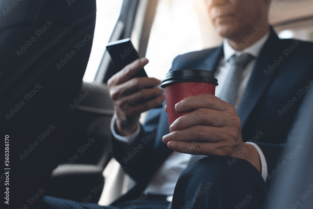 Businessman inside a car on backseat using mobile phone and drinking coffee in the morning Businessman inside a car on backseat using mobile phone and drinking coffee in the morning