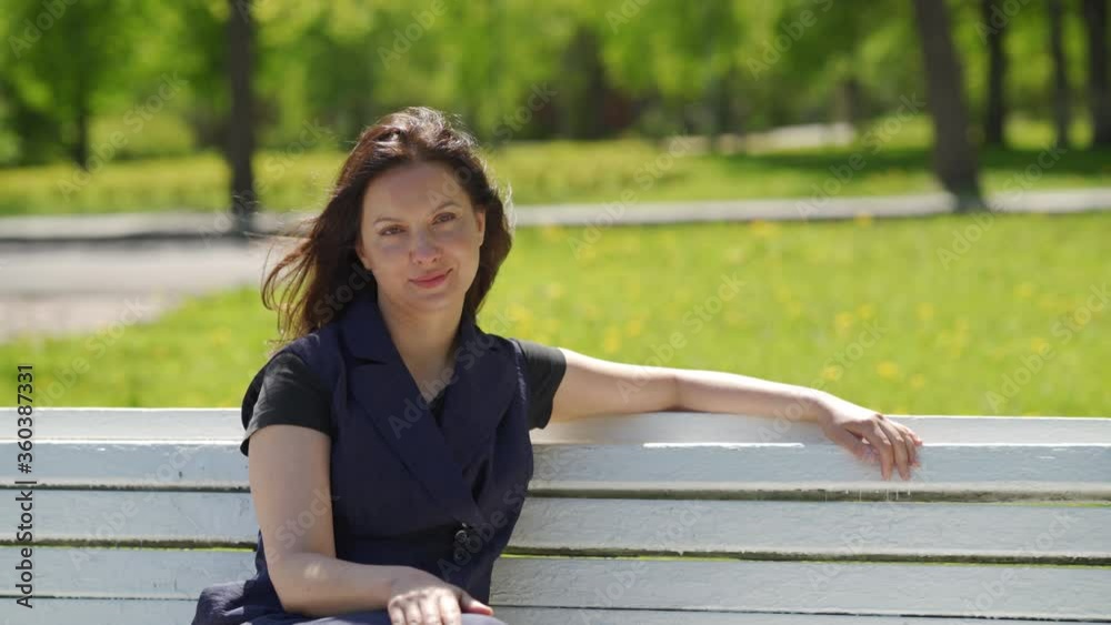 Beautiful brunette woman with long hair sitting on bench in public square on sunny spring day adult woman relaxes in park, looking at camera, smiling. Beautiful brunette woman with long hair sitting on bench in public square on sunny spring day adult woman relaxes in park, looking at camera, smiling.
