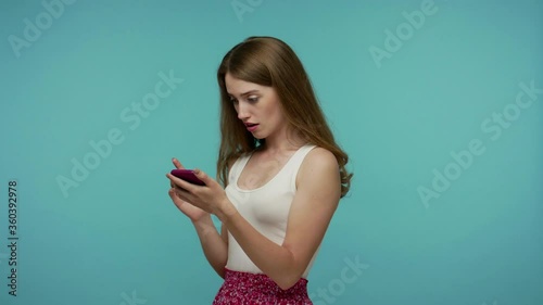 Girl in dress scrolling social networks, found shocking message or image and looking at camera with surprised expression, surfing web using mobile phone . studio shot isolated on blue background photo