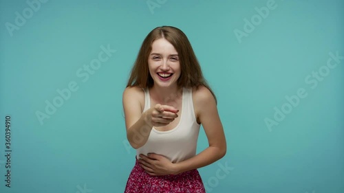 Amused happy girl pointing finger at camera and laughing out loud, holding belly with hilarious laughter, making fun of ridiculous appearance, joke. indoor studio shot isolated on blue background