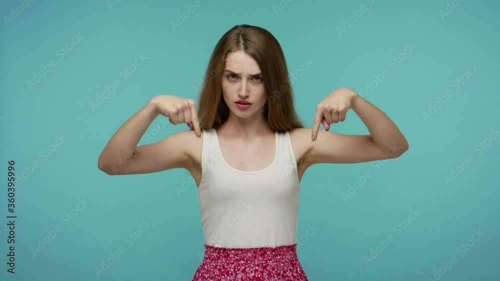Strict girl in dress trying to control, pointing fingers down, demanding immediate act here and right now, looking authoritative, indicating hither. indoor studio shot isolated on blue background Strict girl in dress trying to control, pointing fingers down, demanding immediate act here and right now, looking authoritative, indicating hither. indoor studio shot isolated on blue background