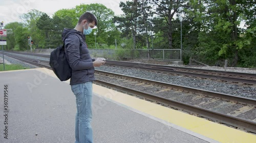 High quality 4k video of a young man who is waiting for a traing on a railway station. GO train station in Canada. Young handsome guy standing on a station and holding his cellphone wearing face mask