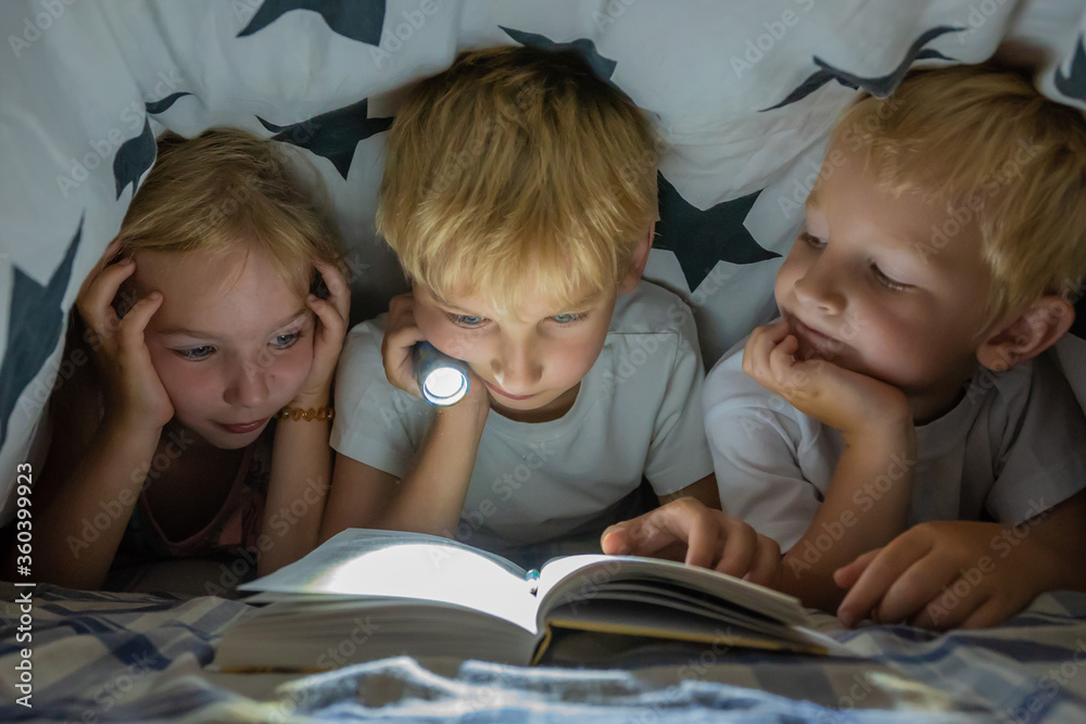 Three young children are reading a book with a flashlight under the covers at night. Three young children are reading a book with a flashlight under the covers at night.