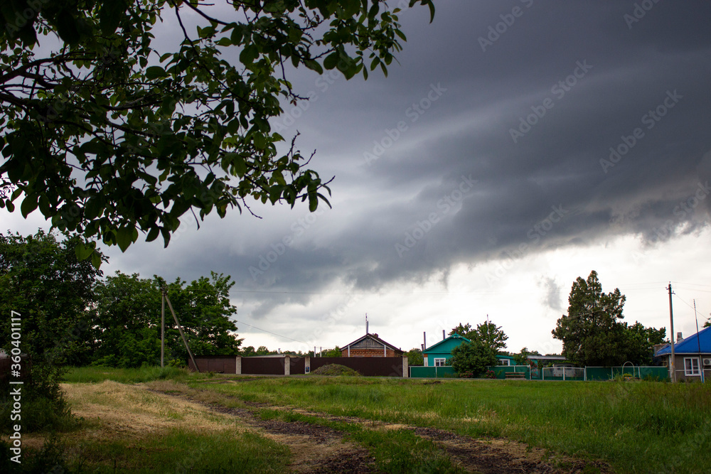 Fototapeta premium Heavy stormy gray sky. Waiting for the rain. Early spring in the village. Green vegetation, trees, bushes, grass. Houses are visible in the distance.