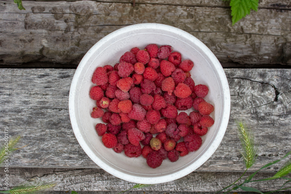 Ripe juicy raspberries in a white bowl. Pink and maroon, berries. On a wooden surface with ears of grass. Ripe juicy raspberries in a white bowl. Pink and maroon, berries. On a wooden surface with ears of grass.