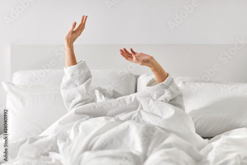 gesture, comfort and morning concept - hands of young woman in hotel robe lying in bed and stretching at bedroom