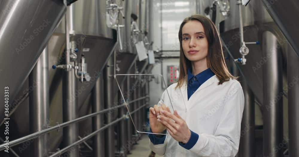Portrait of woman in white lab coat entering data in futuristic transparent glass tablet. Female brewery worker standing at stainless rows of brewing vats and looking to camera Portrait of woman in white lab coat entering data in futuristic transparent glass tablet. Female brewery worker standing at stainless rows of brewing vats and looking to camera