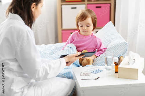 healthcare, medicine and people concept - doctor showing tablet pc computer to sick little girl coughing in bed at home