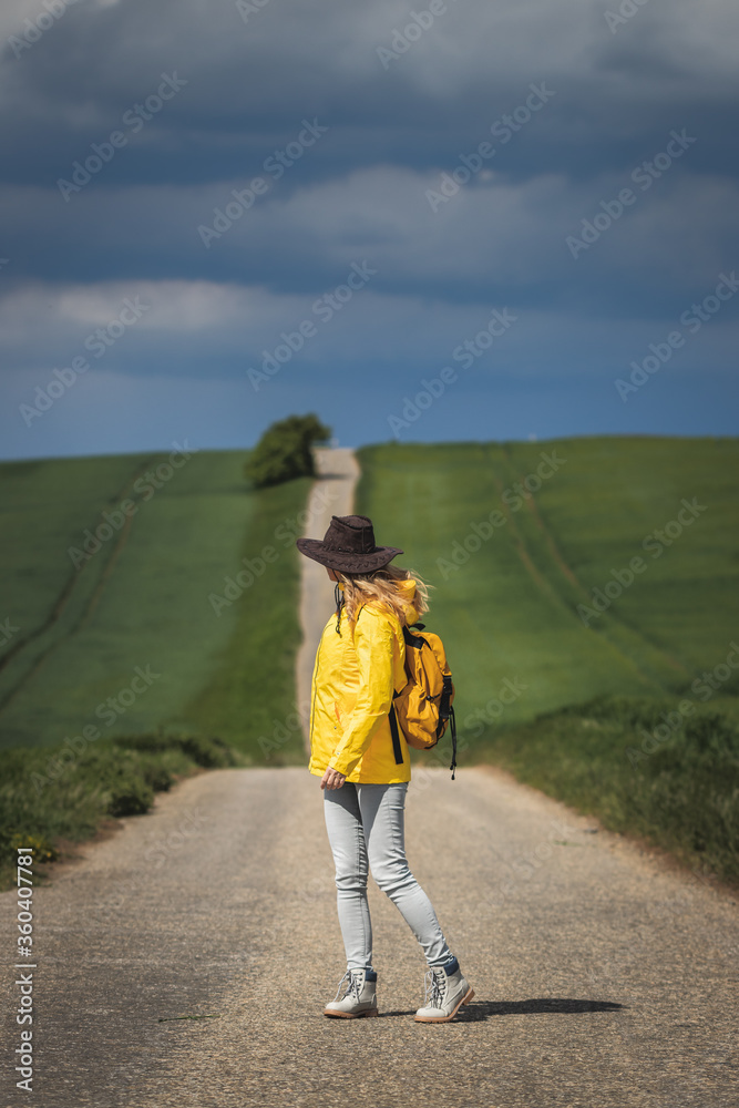 Woman hiking on road and looking at cloudy sky. Rain is coming. Backpacker wearing hat and yellow waterproof jacket	