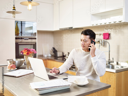 young asian business man working from home looking at laptop computer while making a call to client