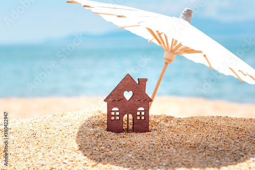 Miniature house and umbrella on beach, blue sea and sky on blurred background. Vacation home for happy holiday for family. Real estate, sale or property investment concept. Closeup. Copy space