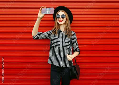 Casual smiling young woman taking selfie picture by smartphone wearing a black round hat, striped shirt on red wall background
