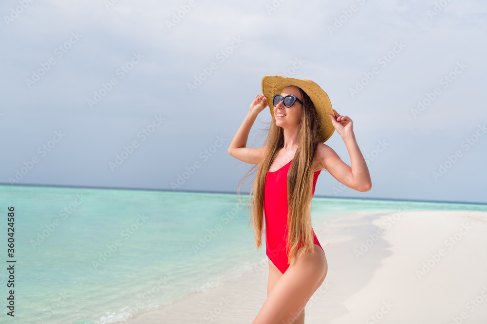Photo of attractive lady long hairdo bronze body skin sunbathing sensual figure skinny shapes seaside resort wear sun headwear red bodysuit enjoy bright sun rays ocean beach outside