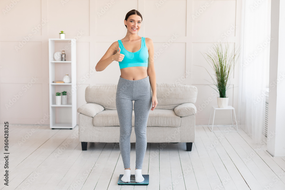 Fit Girl Gesturing Thumbs-Up Standing On Weight Scales At Home Fit Girl Gesturing Thumbs-Up Standing On Weight Scales At Home