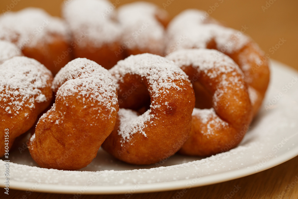 a pile of delicious sweet golden brown doughnuts (donuts) with white powdered icing sugar topping laying in a white plate on a wooden table background