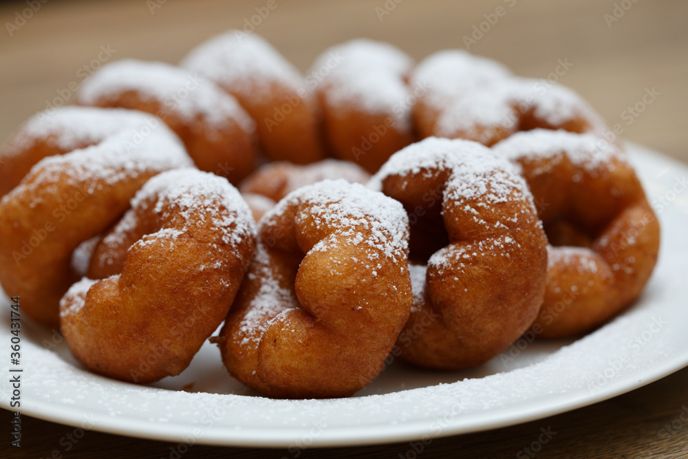a pile of delicious sweet golden brown doughnuts (donuts) with white powdered icing sugar topping laying in a white plate on a wooden table background