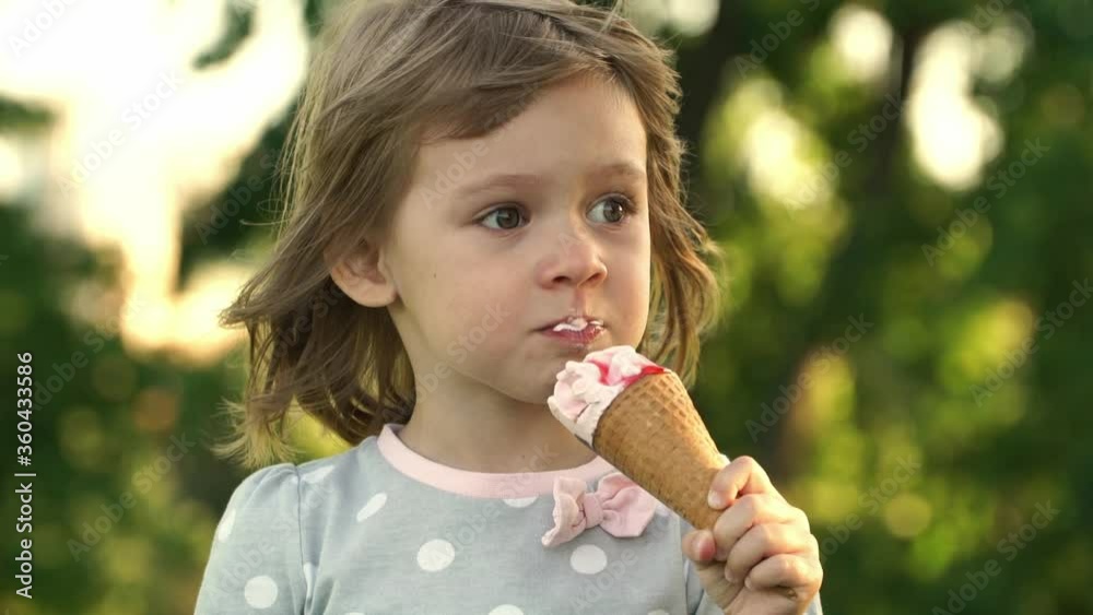 Close up of little happy girl in summer park eats strawberry ice cream cone at sunset on background of lawn, looks at camera and smiles. Child enjoy dessert. Outdoors.