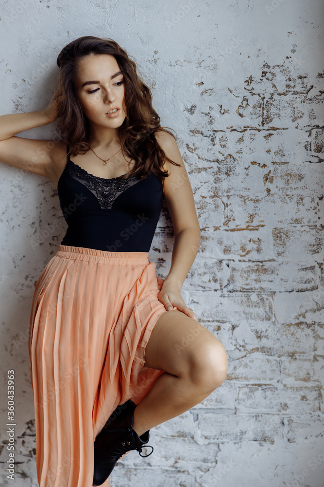 A beautiful brunette girl with curly hair stands against the wall in the loft style. The girl is wearing a light black top and skirt with pleating. High quality photo A beautiful brunette girl with curly hair stands against the wall in the loft style. The girl is wearing a light black top and skirt with pleating. High quality photo