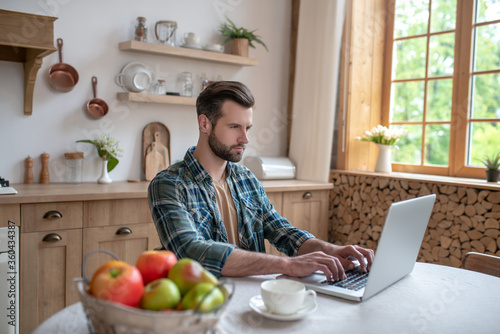 Man in a checkered shirt sitting in the kitchen and working on a laptop
