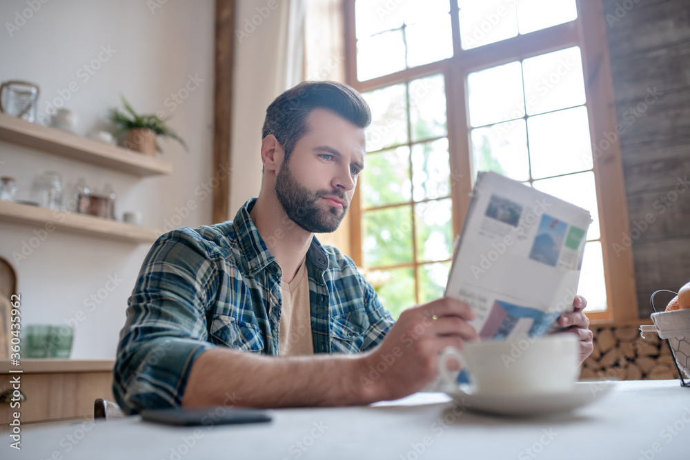 Bearded man in a checkered shirt sitting at the table, reading newspaper and looking interested