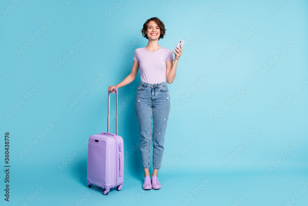 Full size photo of positive cheerful girl tourist hold baggage use smartphone booking taxi hotel wear lilac violet t-shirt denim jeans isolated blue color background