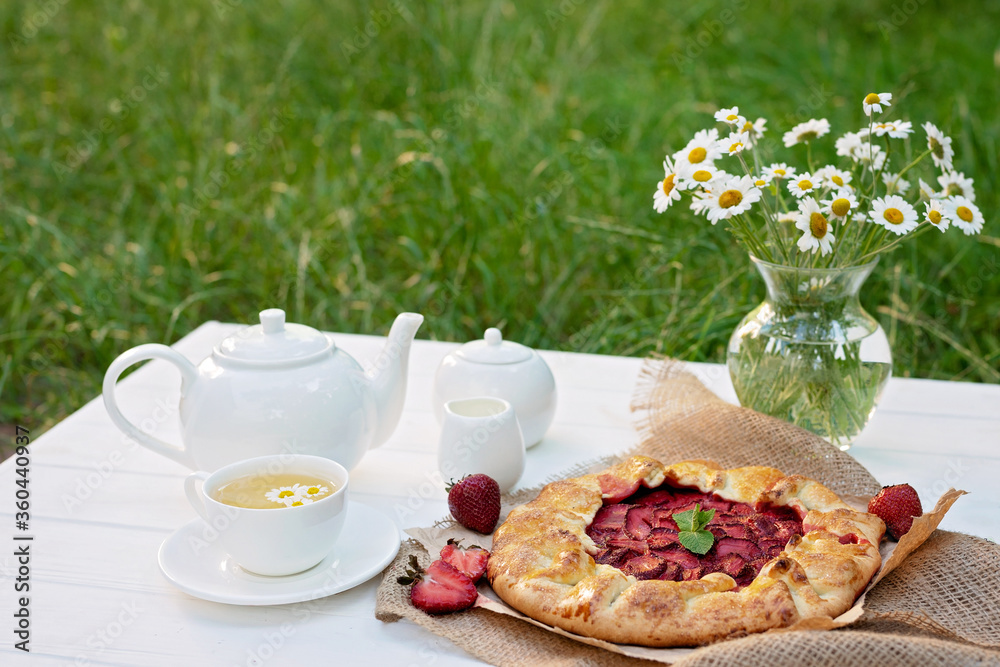 Freshly baked homemade galette or open strawberry pie, a cup of herbal tea, a teapot and a vase with a bouquet of daisy flowers, summer outdoor food. Soft selective focus. Freshly baked homemade galette or open strawberry pie, a cup of herbal tea, a teapot and a vase with a bouquet of daisy flowers, summer outdoor food. Soft selective focus.