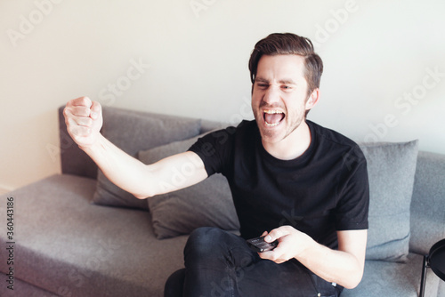 Young handsome man in black t-shirt shaking his head, watching a sport game with emotions. A guy sitting on a sofa, concept of watching TV at home