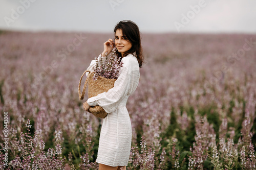 Young brunette woman standing in a big field with pink sage in bloom, holding a wicker bag with flowers. photo