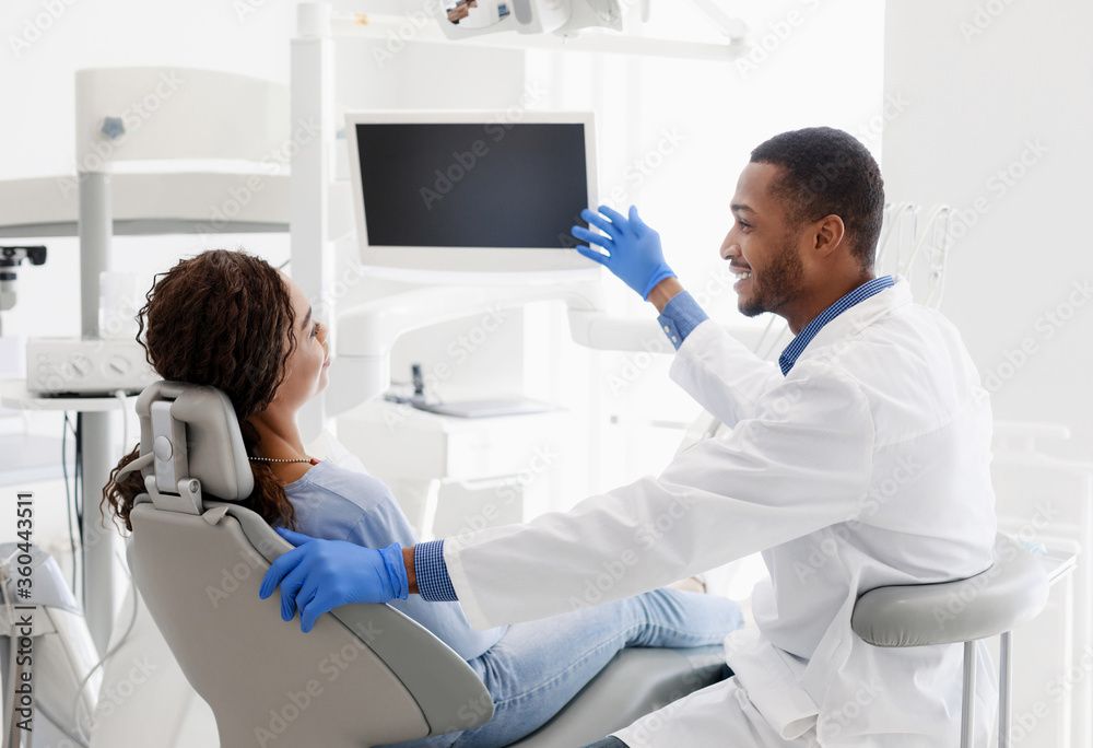 Fototapeta premium African man dentist talking to patient, pointing at blank monitor
