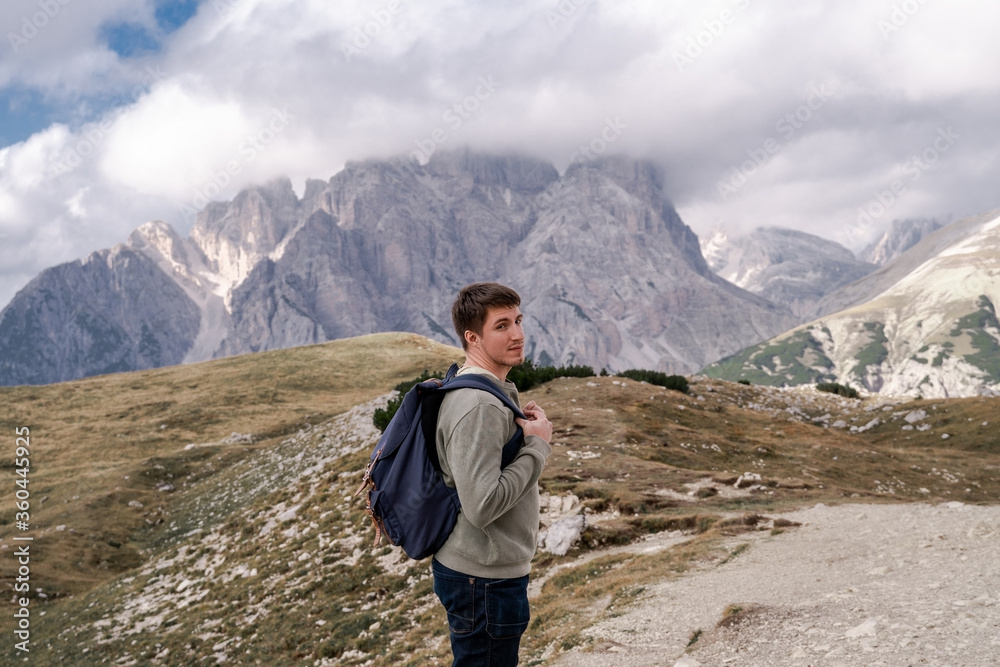 Dolomites Alps. Tre Cime di Lavaredo. Italy. Brunnete hiker with blue backpack walk on alpine trail on background of rocky mountain peaks wrapped by grey clouds in summer