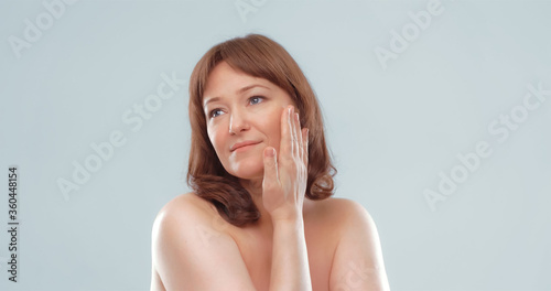 Pretty mature female applies skin care product by touching face with hand. Woman applying anti-age cream on her skin isolated on white background. Spa and wellness concept.