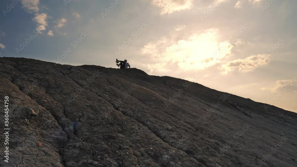 The camera moves up the slope of a rocky hill on the top of which against the background of the sunset sky sits a football freestyler and kicks the ball with his feet, sometimes the athlete kicks the The camera moves up the slope of a rocky hill on the top of which against the background of the sunset sky sits a football freestyler and kicks the ball with his feet, sometimes the athlete kicks the
