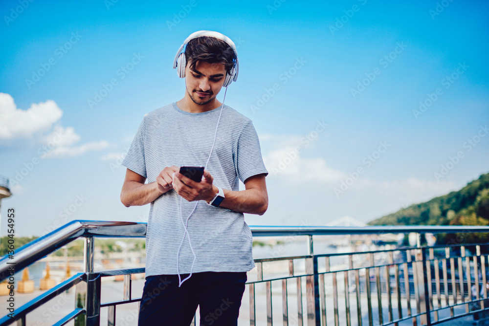 Pensive young man in headphones downloading songs from web page on mobile while spending free time outdoors, hipster guy enjoying music while chatting in social networks with friend using smartphone