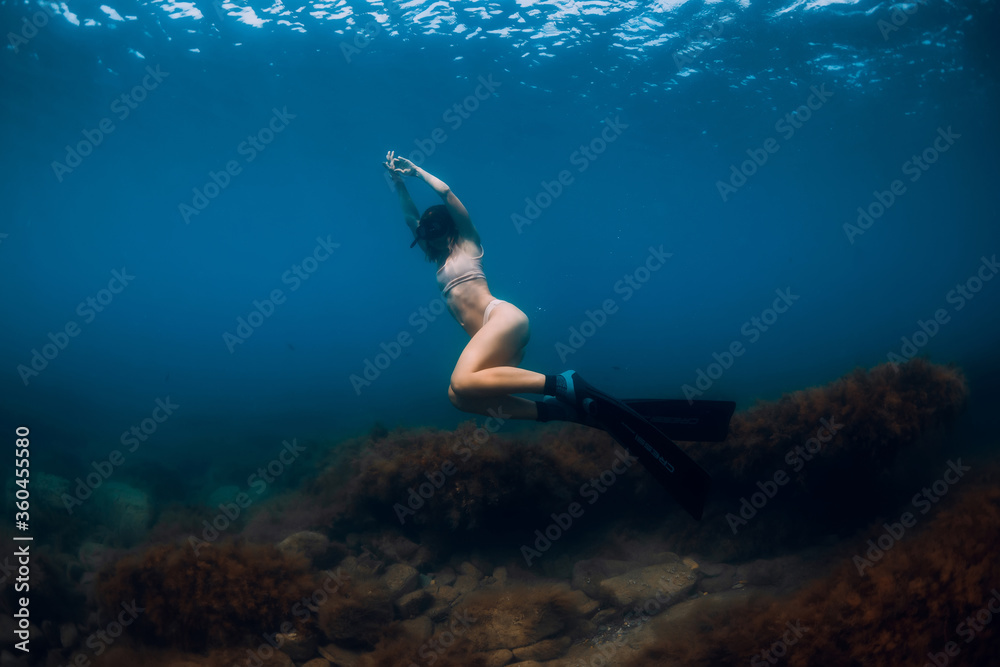 Sporty woman freediver with fins glides underwater in blue sea.