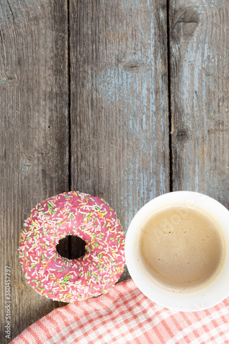 A pink doughnut on a wooden planks background with a cup of coffee with milk and a napkin on a vintage wood background with copy space.