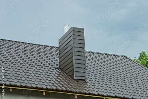 one large metal chimney on a brown tiled roof of a house against a blue sky © butus