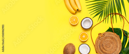 Summer fashion flat lay. Round trendy rattan bag, tropical palm leaves, coconut, orange, bananas on yellow background. Top view, copy space. Trendy bamboo bag. Creative background, vacation concept